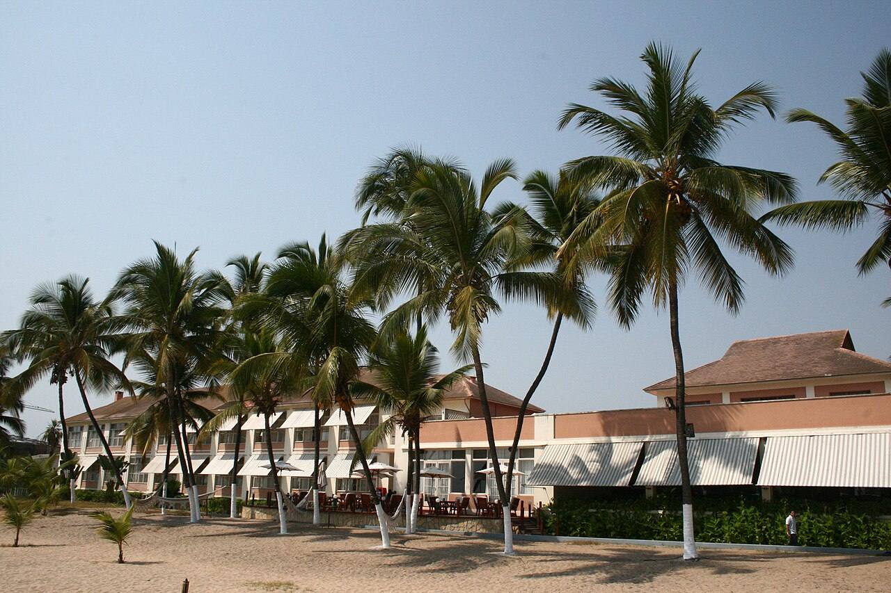 Edifício de hotel com palmeiras e toldos listados numa praia, em Lobito